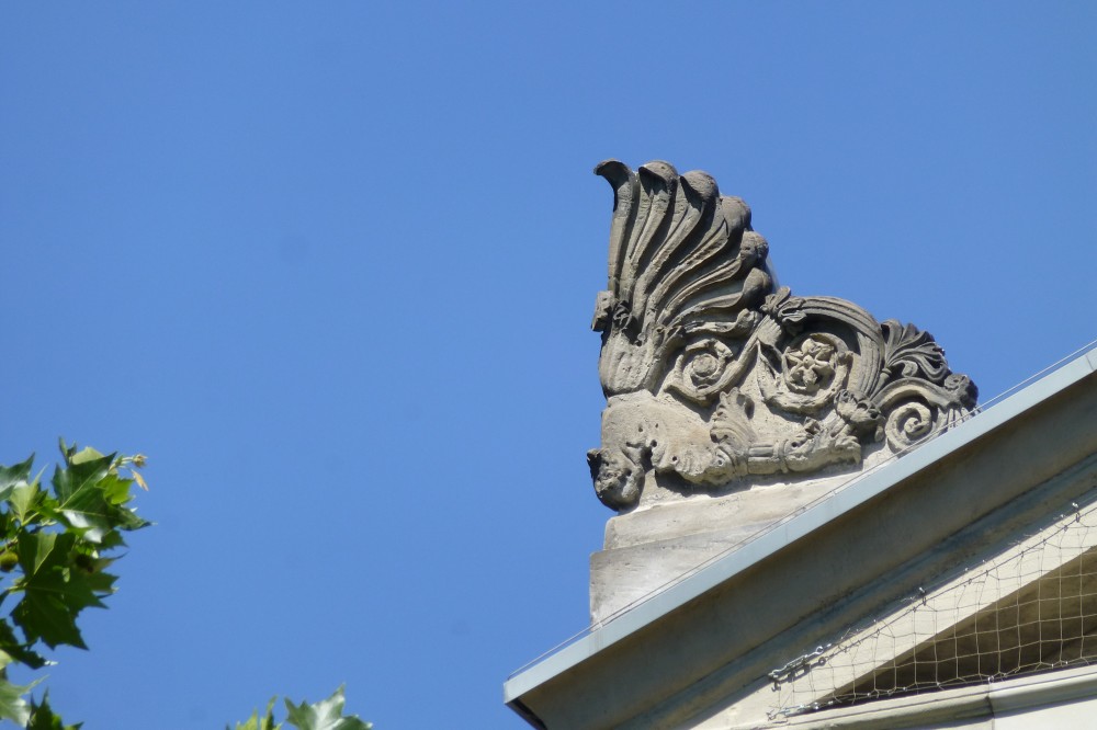 Große steinerne linke seitliche Giebelfigur (Akrotherion) mit Akanthusblättern und anderen floralen Motiven. Der Giebel selbst ist flach geneigt, die Dachkante mit Metall beschlagen, und das Giebelfeld selbst scheint weit zurückzutreten, so dass es im Schatten liegt. Der Hohlraum ist durch ein Taubengitter geschützt. Von links ragen Platanenblätter ins Bild.