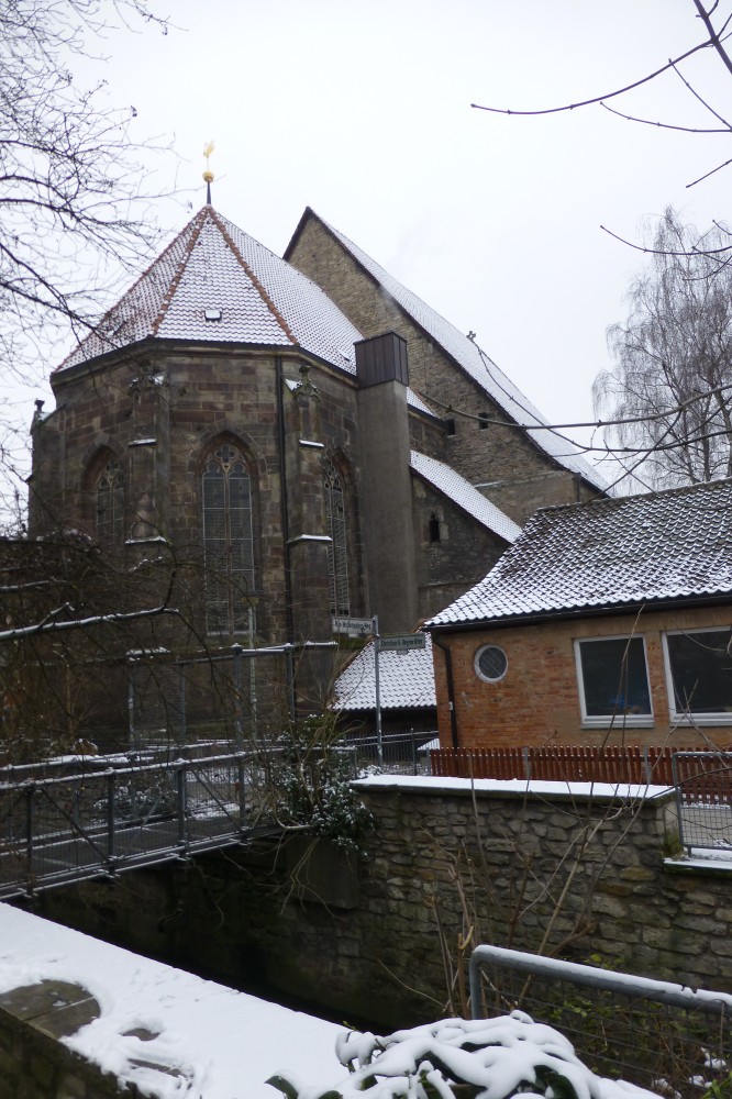 Im Vordergrund ist eine mehrere Meter breite und tiefe Vertiefung mit aus Kalkstein gemauerten Wänden, über den eine kleine Fußgängerbrücke aus Metall führt. Was sich auf dem Grund befindet, ist nicht zu sehen, womöglich ist es ein Wasserkanal oder eine alte kleine Bahnlinie. Dahinter ragt von links ein ebenerdiges Ziegelhaus mit zwei fast quadratischen und einem kleinen runden Fenster ins Bild, davor ein rotbraun angestrichener Holzzaun. Rechts steht ein großes kirchenartiges Gebäude mit spitzem Satteldach, daran angebaut ein etwas niedrigerer Altarraum mit Spitzbogenfenstern. Dieser hat wiederum zwei kleinere Anbauten mit einem hohen Schornstein, der bis über die Dachkante des Altarraums reicht. Die Bäume sind kahl, und es liegt etwas Schnee.