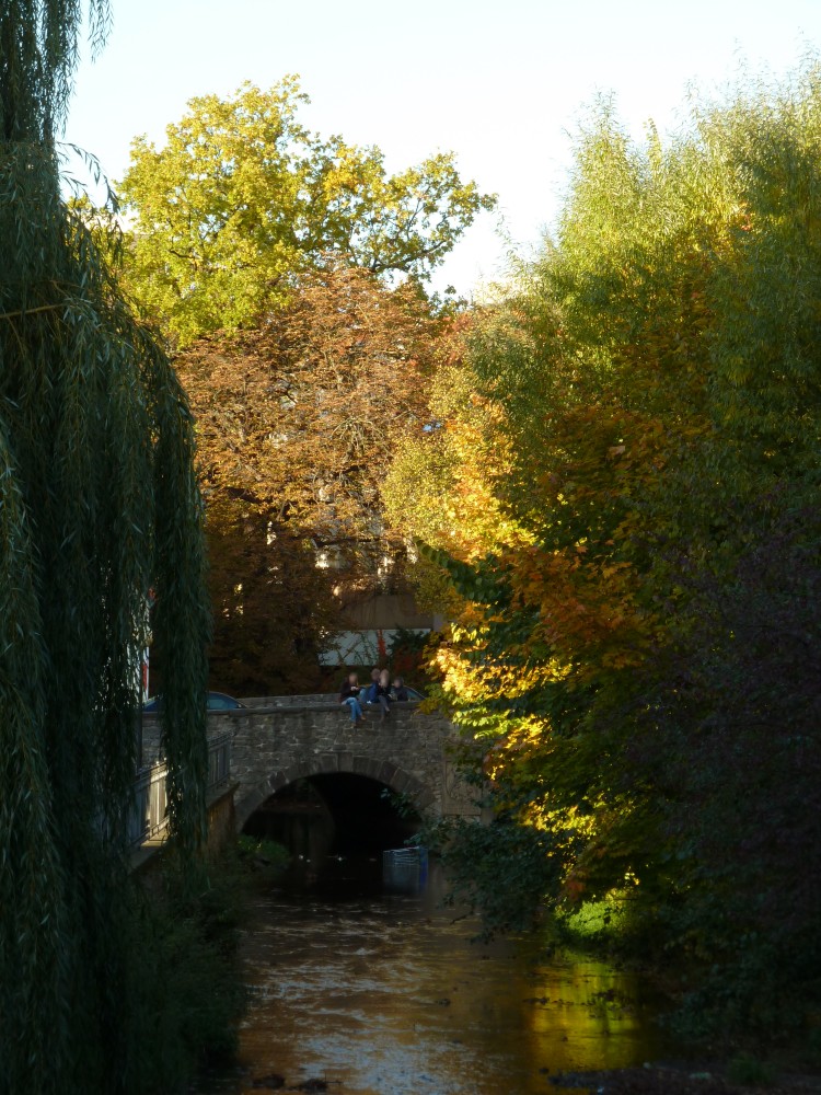 Man sieht einen Wasserlauf mit dichten frühherbstlichen Bäumen an beiden Ufern. Durch das Laub sieht man im Hintergrund eine gemauerte Bogenbrücke, von der ein Bogen vollständig zu sehen und ein zweiter zu erahnen ist. Auf der Brückenmauer sitzen Menschen. Über die Brücke fährt ein Auto. Dahinter sind Häuser zu sehen.