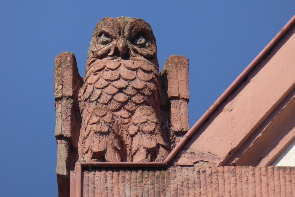 Vor dem blauen Himmel sieht man eine hell rotbraune Eulenfigur, deren Flügel sehr rechteckig aussehen. Die Federn an Brust und Bauch sind wie Schuppen dargestellt. Es sieht aus, als säße die Eule auf der flachen Ecke eines Giebels, der erst rechts von der Eule schräg nach oben führt. Die sichtbaren Bauteile haben dieselbe rötliche Farbe wie die Eule.
