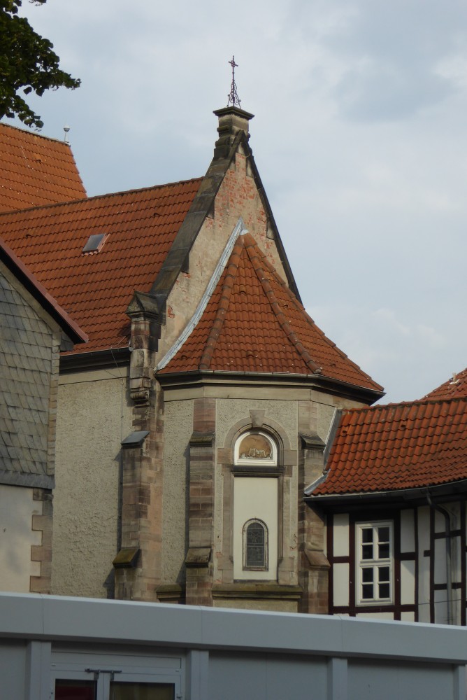 Im Vordergrund verläuft der obere Teil eines flachen Metallbaues quer durch das Bild. Dahinter ragt von links ein verputztes Steinhaus ins Bild, dessen Ecke in sichtbarem Sandstein ausgeführt ist und dessen Giebel mit Schiefer oder Schieferimitat behängt ist. Dahinter ebenfalls von links bis in die Bildmitte ein hoher, ebenfalls verputzter Steinbau mit Strebepfeilern und erhöhter Giebelmauer, auf deren Spitze sich ein metallenes Kreuz befindet. An den Giebel ist ein Chor angebaut, der ein teilweise verdunkeltes Rundbogenfenster hat. Von rechts stößt ein etwas niedrigerer Fachwerkbau an den Chor.