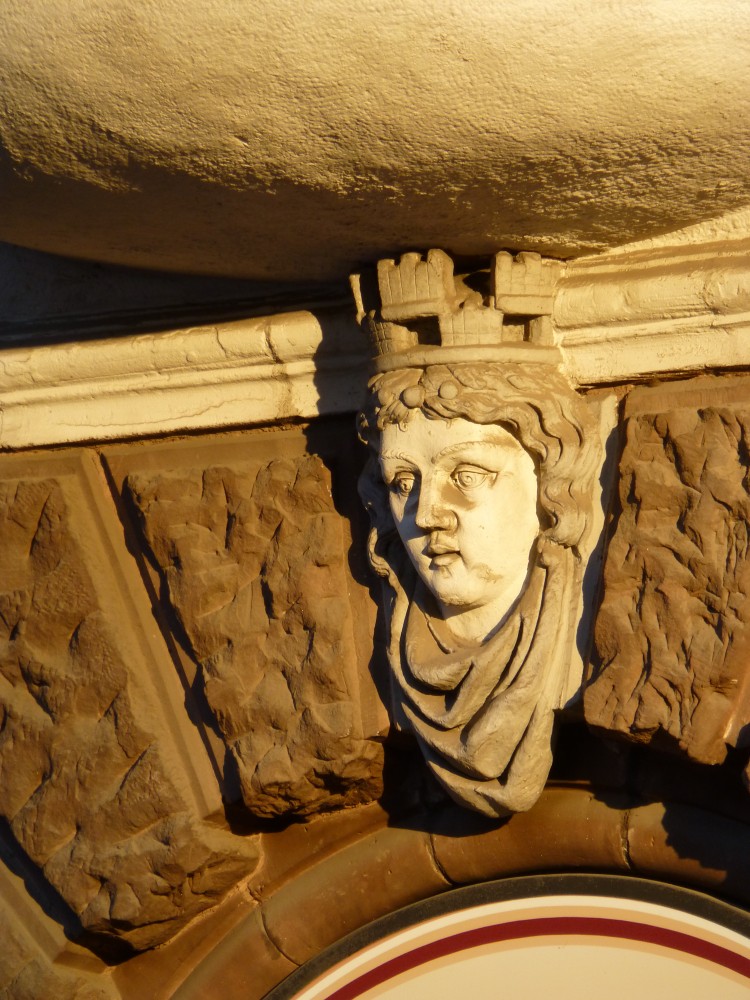 Das Foto enthält vier Steine aus dem Bogen über einer Tür. Drei davon sind behauene Sandsteine mit absichtlich unebener, grob strukturierter Oberfläche. Der oberste Stein ist die plastische Darstellung eines Gesichtes mit welliger Haarfrisur, faltigem Halstuch und einer Art doppelter Krone. Unter der Krone ist ein weiterer Reif mit drei Steinen angedeutet. Der Mund ist sehr klein, die Nase gerade, die Augenbrauenbogen groß und geschwungen. 
				Haar und Krone reichen über den Abschluss des Türbogens hinaus bis unter eine gebogene verputzte Fläche, die wahrscheinlich die Unterseite eines Balkons oder Erkers darstellt. 
				Unterhalb des Türbogens sieht man einen Teil der Tür oder des Rahmens mit mehreren Streifen entlang des Bogens, von denen ein Streifen auffällig dunkelrot ist.