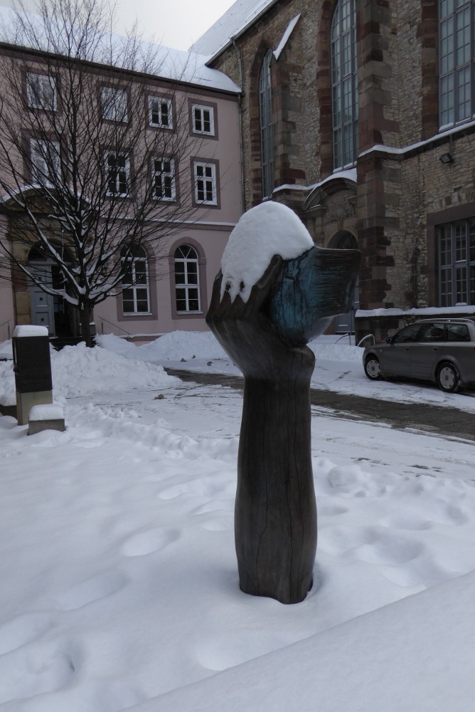 Bronzene Figur einer nach oben gestreckten Hand, die einen Fisch hält, in einem verschneiten Innenhof; ganz links im Mittelgrund ein schwarzer Kasten, etwa doppelt so hoch wie breit, auf einem grauen Sockel. Dahinter ein Barockgebäude, das im rechten Winkel an ein rechts im Bild befindliches gotisches Kirchenschiff mit Barockportal angebaut ist. Ganz rechts im Mittelgrund steht ein graues Auto.