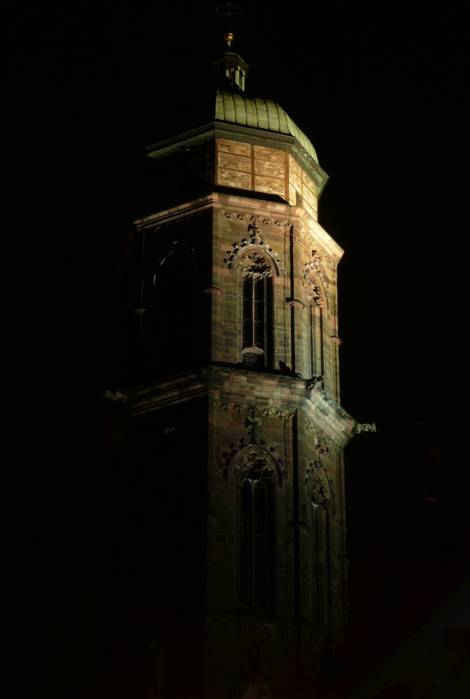 Angestrahlter Kirchturm vor einem schwarzen Nachthimmel. Man sieht drei der wahrscheinlich acht Kanten des Turmes. Der Turm hat drei Simse, zwischen denen gotische Fenster angeordnet sind. Das mittlere Sims ist am breitesten und hat Wasserspeier an den Ecken. Über dem obersten Sims erhebt sich ein einfacher Aufbau aus ausgemauertem Fachwerk, darüber eine Welsche Haube, die mit bis zur Grünfärbung korrodiertem Kupferblech gedeckt ist. Ganz oben lässt ein glänzender Lichtpunkt eine goldfarbene Zeitkapsel erahnen.