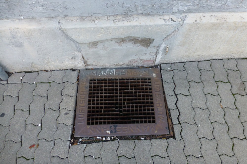 Im oberen Teil des Bildes sieht man den verputzten Sockel eines Hauses, darunter eine mit UNI-Verbundpflaster gepflasterte Fläche. In diese Fläche ist, an den Haussockel angrenzend, ein quadratisches Metallgitter mit breitem Rand eingelassen. Auf dem Rand steht in Reliefbuchstaben: MANNESMANN und ein Wort, das mit Z aufhört. Direkt über dem Gitter weist der Haussockel Risse auf.