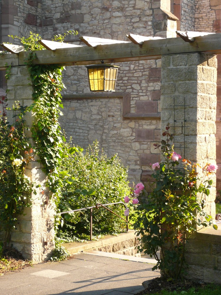 Durchgang zwischen zwei gemauerten Steinsäulen, die oben durch eine hölzerne Pergola verbunden sind, teilweise mit Rosen bewachsen. In der Mitte der Pergola hängt eine gelbe Laterne. Direkt hinter dem Durchgang führt eine Treppe nach unten. Im Hintergrund die Wand eines Gebäudes, teilweise aus behauenem Sandstein und teilweise aus Kalkstein.