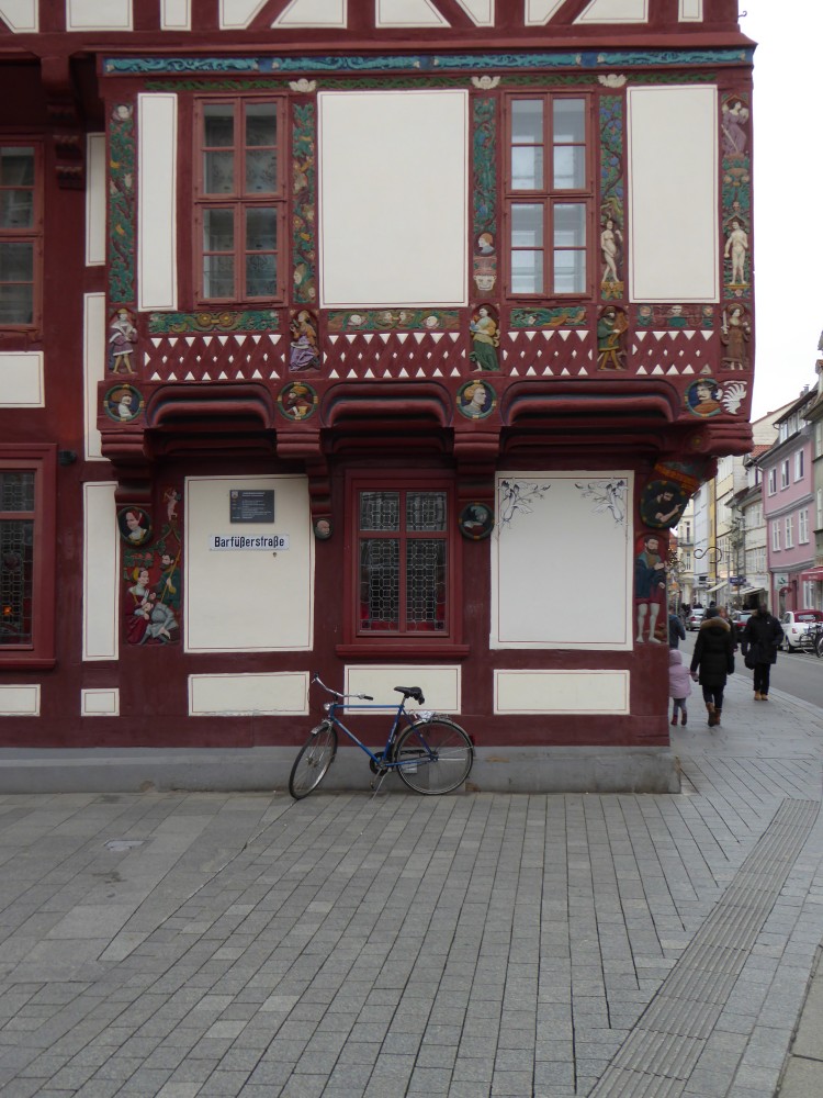 Man sieht Erdgeschoss und Zwischengeschoss eines Eckhauses. Die Ecke ist links im Bild. Das Zwischengeschoss ist um die Ecke herum als Erker herausgebaut. Am und unter dem Erker sind viele farbige figürliche Schnitzereien an allen Balken: menschiche Figuren, runde Medaillons mit Köpfen, grüne Ranken, in die ebenfalls Gesichter eingearbeitet sind. Das Holz, soweit nicht andersfarbig bemalt, ist rotbraun, die Gefache hellbeige. Am Rand jedes Gefachs verläuft eine feine rotbraune Linie, das rechte untere hat außerdem feine schwarze aufgemalte Ornamente in den oberen Ecken. Am gesamten Erker sind die unteren Gefache mit schrägen, sich kreuzenden Hölzern fast vollständig ausgefüllt. Vor dem Haus steht ein Fahrrad. In der Straße rechts sind mehrere Fußgänger und parkende Autos zu sehen.