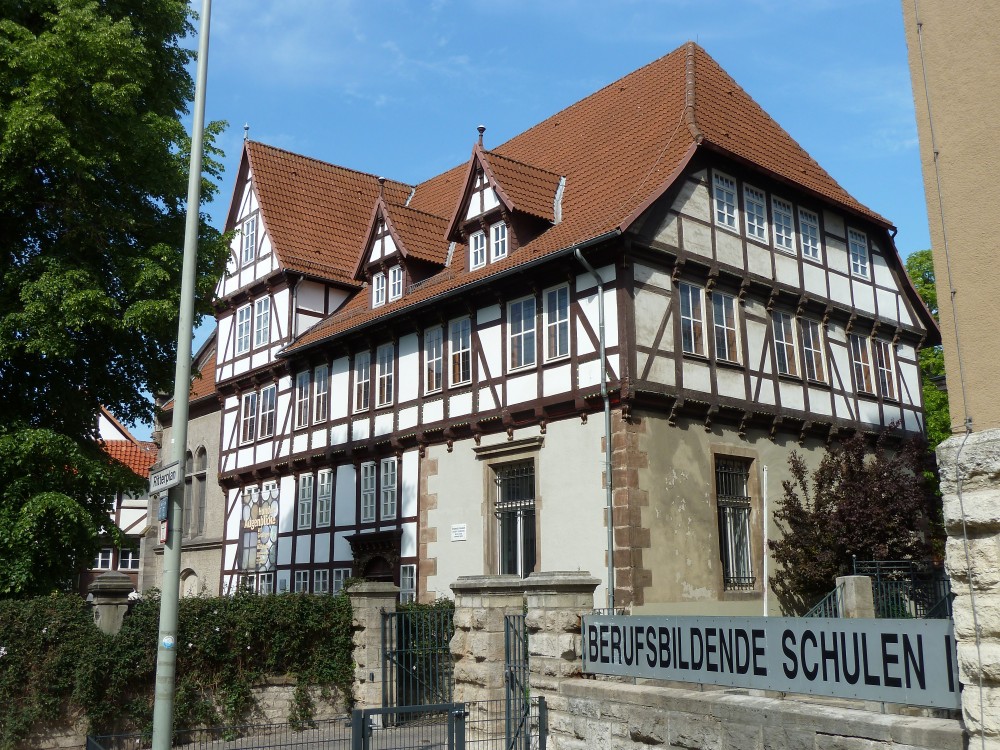 Fachwerkpalais, das mit einigen Metern Abstand zur Straße frei steht. Rechts unten ist ein Teil des Hauses kein Fachwerk, sondern Stein, und zwar verputzt mit freiliegenden behauenen Sandsteinen um die Fenster und an der Hausecke. Der Steinteil nimmt eingeschossig die Höhe von zwei Fachwerkgeschossen ein. Neben dem Steinteil hat das Palais ein hölzernes reich verziertes Rundbogenportal. Das Oberstockwerk steht auf beiden sichtbaren Seiten etwas über (Vorkragung) und hat auf der Straßenseite fünfmal zwei und auf der Schmalseite dreimal zwei Fenster. Vor dem ganz linken Fensterpaar hängt ein Banner. Die Vorkragung ist mit Schnitzereien geschmückt. Auf der Straßenseite beginnt über dem Oberstockwerk das Dach, auf der Schmalseite noch ein Stockwerk höher (Krüppelwalmdach). Das Dach auf der Straßenseite hat einen Scheingiebel ganz links sowie zwei kleinere Gauben.
				Links hat das Palais einen steinernen Anbau mit Rundogenfenstern. 
				Im Vorgarten steht eine große Linde. Durch die Zweige blickt man auf ein weiteres Fachwerkhaus links.
				Der Vorgarten ist durch eine Kombination aus Mauer (unten) und üppig beranktem Zaun (oben) von der Straße getrennt. Große Steinpfeiler bilden die Zaunpfähle. Rechts davon schließt sich mit weiteren Pfeilern eine Einfahrt mit Gittertor und eine Gitterpforte für Fußgänger an. Danach geht die niedrige Gartenmauer weiter bis ans Nachbarhaus, das an die Straße gebaut ist und dessen Ecke gerade noch ganz rechts im Bild zu sehen ist. Anstatt eines begrünten Zaunes gibt es hier oberhalb der Mauer aber ein Schild: Berufsbildende Schulen II. 