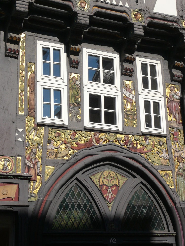 Man sieht den oberen Teil eines spitzen Torbogens und die darüber befindliche Wand bis zur Vorkragung des Oberstockwerks. Links ragt der Erker ins Bild. Die Wand über dem Torbogen ist fast vollständig mit Schnitzereien bedeckt: Personen, Fabelwesen, Ranken mit eingearbeiteten Gesichtern. Eine Person hält ein Spruchband mit der Zahl fünfzehnhundertneunundvierzig. Die Wand hat drei Fenster, die offenbar später eingebaut worden sind, da das linke Fenster Schnitzfiguren abschneidet. Der Torbogen ist später in zwei Türbögen aufgeteilt worden, zwischen denen farblich zum älteren Schnitzwerk passend ein Zirkel, weiteres Werkzeug sowie stilisiertes Blattwerk abgebildet ist. 
				Die Gefache - nur am Oberstockwerk erkennbar - sind weiß, das Holz dort, wo keine Schnitzereien sind, dunkelbraun.