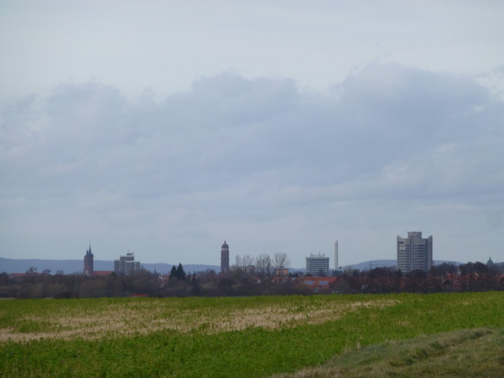 Blick über ein winterliches, aber grünes Feld hin zur Stadt Göttingen. Von links nach rechts sind die markantesten Bauten: 1. eine Kirche mit zwei hohen, aber ungleich hohen spitzen Türmen, wobei die Türme auf der linken Seite sind, 2. ein ziegelrotes Wohn-Hochhaus aus mehreren Blöcken, 3. direkt rechts davon eine weitere Turmspitze, die aber niedrig und unauffällig ist, 4. ein übermäßig hoher Kirchturm mit Welscher Haube, 5. ein nur mittelhoher Beton-Glas-Block mit abgeschrägten Ecken, 6. direkt rechts davon ein sehr hoher weißer doppelter Schornstein, 7. ein hohes mehreckiges zusammengesetztes Beton-Glas-Hochhaus mit hochgezogenen Außenkanten und erhöhtem Mittelbau - dieses Gebäude scheint dem Betrachter am nächsten zu stehen - und schließlich 8. am rechten Bildrand eine weitere Welsche Haube, die aber kaum über den Horizont ragt. Das gesamte Gelände fällt nach links leicht ab.