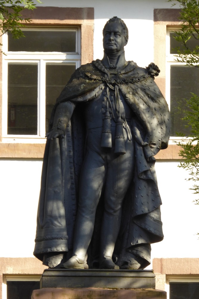 Bronzestatue eines Mannes in einem weiten geschmückten Umhang, der vorn offen ist, so dass darunter eng anliegende Kleidung zu sehen ist. Vor dem Bauch hängen zwei konische Gegenstände herab. Auf der linken Schulter ist eine Art Rosenblüte befestigt. Aus dem Kragen des Umhangs ragt ein weiterer, steifer, den Hals bis fast oben umfangender Kragen hervor. Der Mann trägt weder Bart noch Kopfbedeckung. Im Hintergrund ist ein verputztes Gebäude zu sehen, dessen Fenster mit sichtbarem Sandstein eingefasst sind. Zwei Fenster befinden sich auf der Höhe des außerhalb des Bildes befindlichen Sockels, so dass nur deren oberes Ende im Bild zu sehen ist. Zwei weitere Fenster reichen vom Bauch bis zum Kopf der Skulptur. Von der Seite ragen grüne Zweige ins Bild. Auch das fleckige Sonnenlicht auf der Skulptur deutet auf die Nachbarschaft von Bäumen hin.