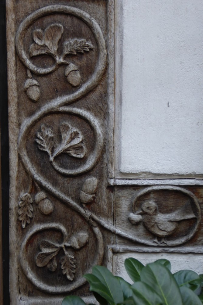 Detail eines senkrechten Fachwerkbalkens, in den gewundene Ranken mit Eichenlaub, anderem Laub und Eicheln geschnitzt sind. Eine Ranke reicht in einen Querbalken hinein und auf dieser sitzt ein Vogel, der einen herzförmigen Gegenstand im Schnabel hält. Das Holz ist dunkel holzfarben, die Gefache weiß. Von unten ragen glänzende dunkelgrüne Blätter ins Bild.