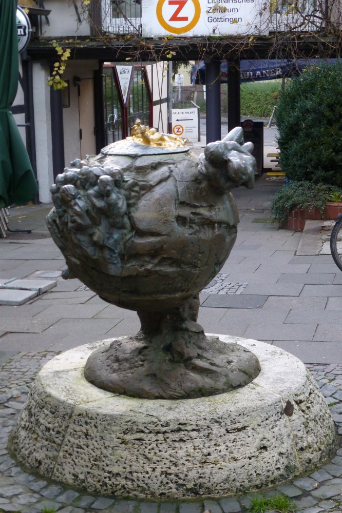 Bronzeskulptur in Form einer teilweise eingerissenen Kugel mit Menschen in verschieden Größen und Stellungen darauf. Im Hintergrund sieht man einen Durchgang, eine recht modern aussehende Fachwerkfassade links und ein großes Banner mit einem roten Z in einem orangen Kreis und der Aufschrift: Literarisches Zentrum Göttingen, direkt über dem Durchgang. Das gleiche steht hinter dem Durchgang noch einmal auf einem Aufstellschild mit einem Pfeil nach links. 