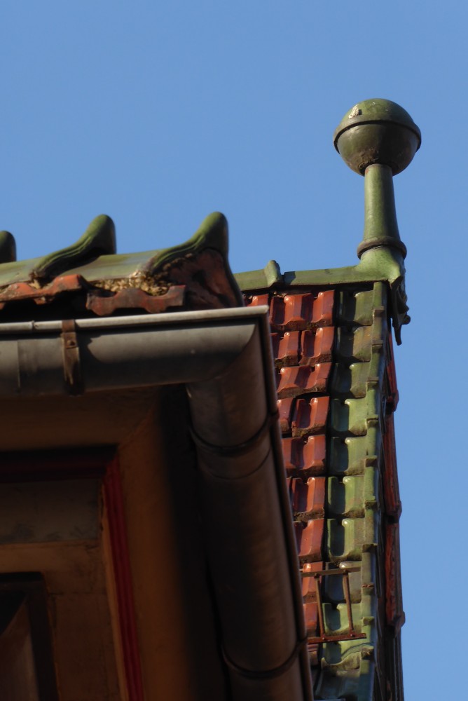 Man blickt von schräg unten unter die Ecke eines Daches, das offenbar ein Walmdach ist und rechts ein Zwerchhaus (einen kleinen Giebel) hat. Die Ziegel auf der Dachfläche des Zwerchhauses (soweit sichtbar) sind glänzend rot, ebenso wie die Ziegel direkt an der Dachrinne. Die Ziegel an der Kante des Giebels und die Firstziegel sind glänzend grün. Jeder Firstziegel hat eine hochgezogene geschwungene Spitze. Der Endziegel auf der Spitze des Giebels hat eine Fläche, die über die Giebelseite herunterragt und offenbar ein Relief aufweist, und eine sehr hochgezogene Spitze, die oben in eine ebenfalls grüne Kugel mündet. Die Kugel hat eine stark abgesetzte Verdickung um den Äquator, so dass man aud die Idee kommt, es könnte sich um eine Zeitkapsel handeln.