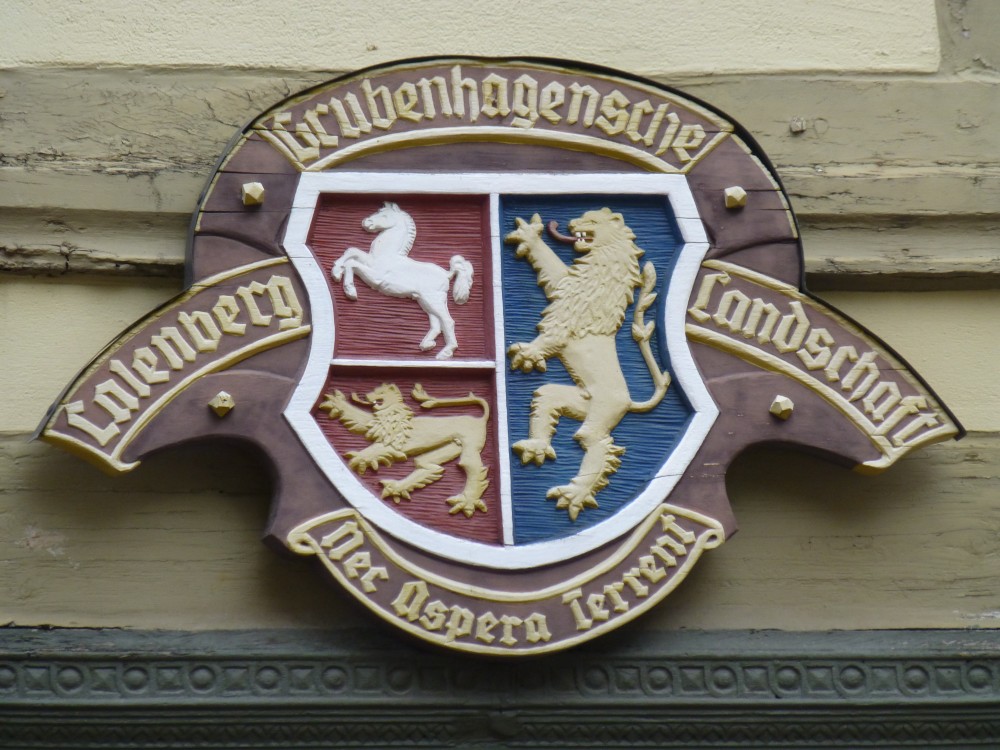 Hölzernes, an einer Fachwerkfassade angebrachtes Schild. In der Mitte ist ein Wappen, darum herum golden eingerahmte Spruchbänder mit goldener Reliefschrift: links: Calenberg, oben: Grubenhagensche, rechts: Landschaft, unten: Nec Aspera Terrent. Das Wappen selbst ist senkrecht geteilt und die linke Hälfte noch einmal waagerecht geteilt. Die beiden linken Felder haben einen roten Hintergrund. Im oberen ist ein weißes Niedersachsenross als Relief dargestellt, im unteren ein goldenes löwenartiges Tier, das nach links schaut und auf allen Vieren nach links läuft. Das rechte Feld ist blaugrundig und entählt ebenfalls ein goldenes löwenartiges Tier, das nach links gewandt ist, dieses steht aber auf den Hinterbeinen.
				Die Fachwerkfassade hat hell olivgrüne Balken und noch hellere ebenfalls grünliche Gefache. Am unteren Bildrand ist eine dunkelgraue beschnitzte Leiste zu sehen, wahrschenlich ein Türrahmen.